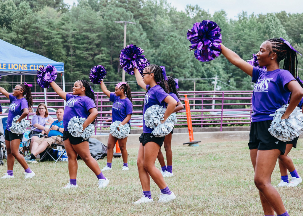 Cheerleaders wearing purple shirts and black shorts hold one pom pom in the air, and the other on their hip as they perform a cheer on a grassy field outside.