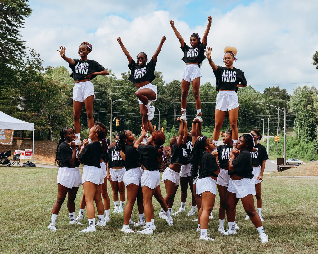 Cheerleaders stand in a grassy field, performing a stunt. Several stunt groups hold cheerleaders in the air.