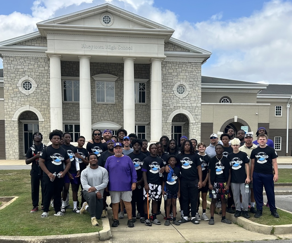 A group of football players wearing matching shirts stand next to coaching staff in front of Hueytown High School