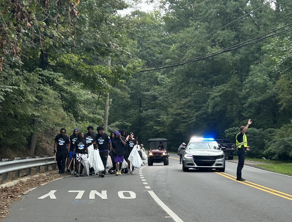 A group of football players wearing matching shirts hold trash bags and pickup trash alongside a road. A police officer stands in the middle of the road, directing traffic.