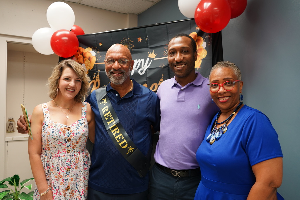 Two men and two women smile and stand together for a photo. They stand in front of a banner with balloons on it.