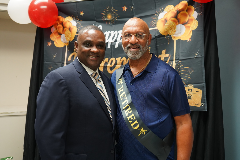 Two men stand together in front of a banner with balloons on it and smile. One of the men wears a sash that says "retired"
