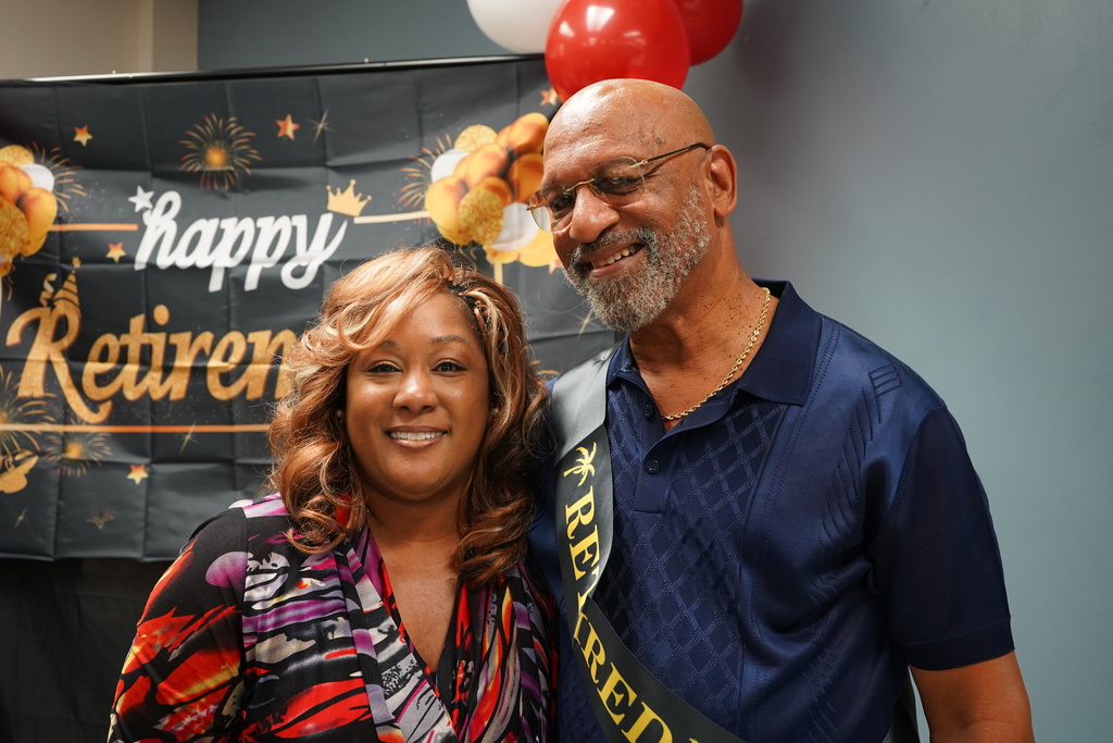 A woman and man smile and stand together for a photo. They're standing in front of a sign that says "Happy Retirement."