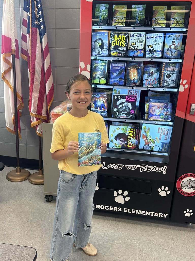 A girl smiles while holding a book and standing in front of a book vending machine