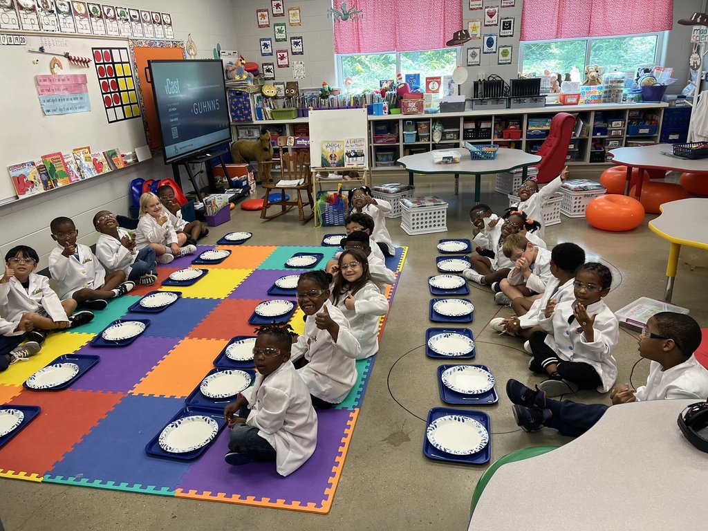 Students wearing lab coats sit on the floor of a classroom. Each student has a tray with a paper plate on it sitting in front of them.