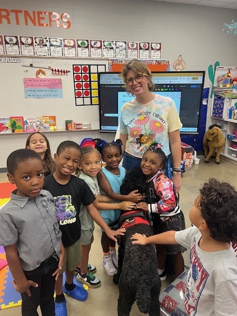 Students and a counselor stand next to a therapy dog in a classroom. Some of the students hug the dog.