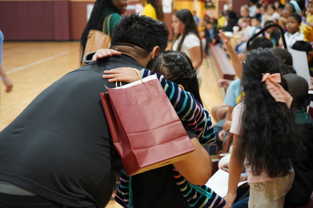 A man and a little girl hug while standing in a school gym.