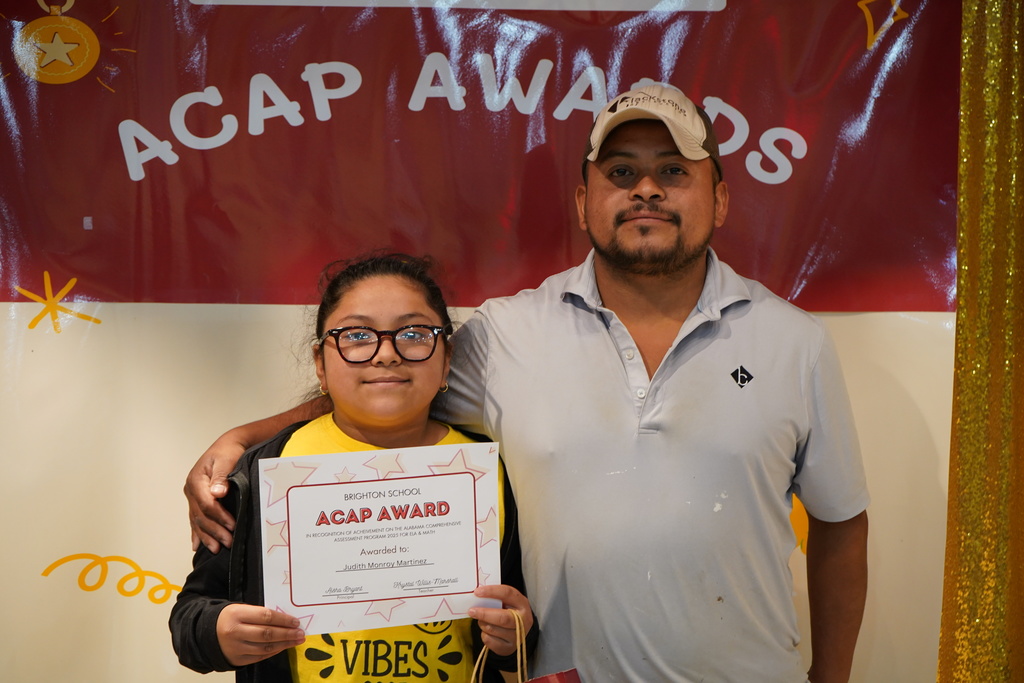 A girl and a man stand together and smile. The little girl is holding an award.