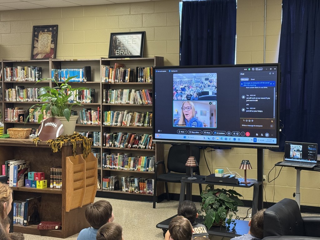 Students sit in front of a monitor in a library. The screen shows a video call between the students and a woman