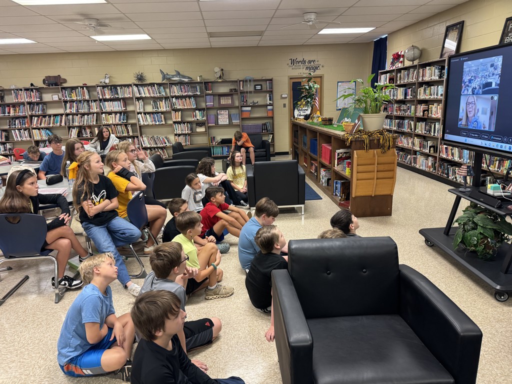 Students sit in a library and stare at a monitor. The monitor shows a video call between the students and a woman.