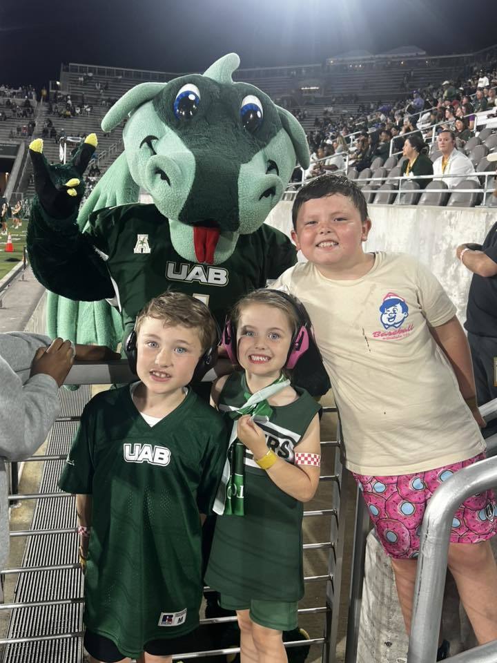 Elementary-aged students smile while standing next to the Blaze mascot at a UAB Football game