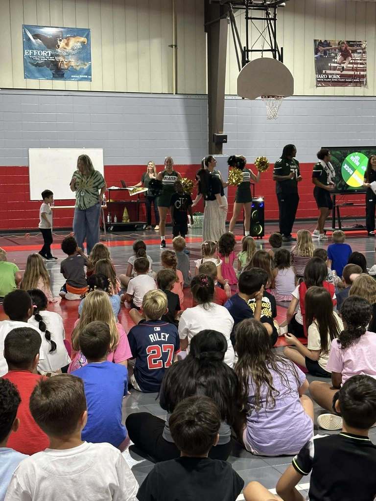 Elementary-aged students sit on the floor of a gym while cheerleaders and football players stand in front of them