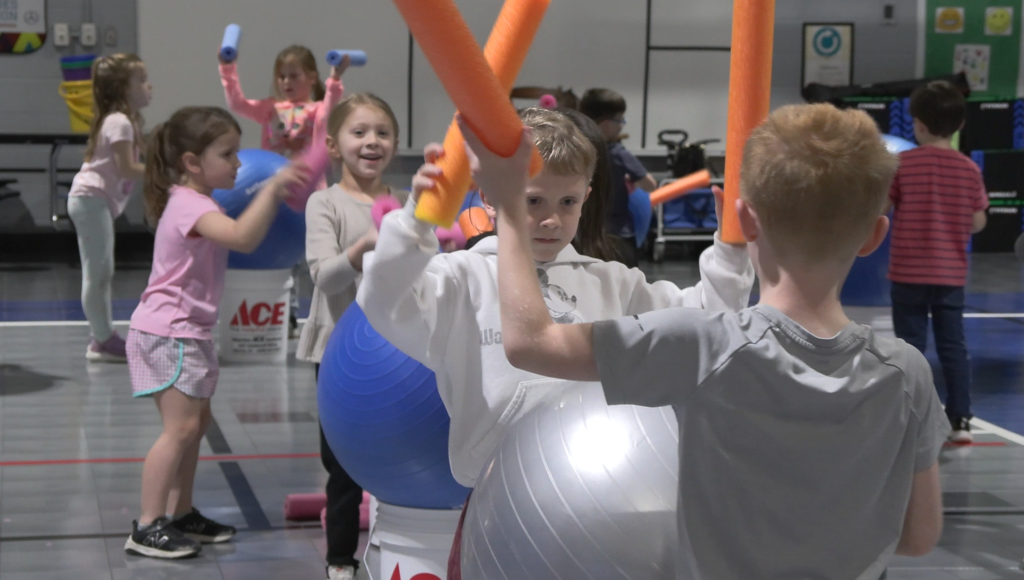 Children hit pool noodles on yoga balls inside a gym.