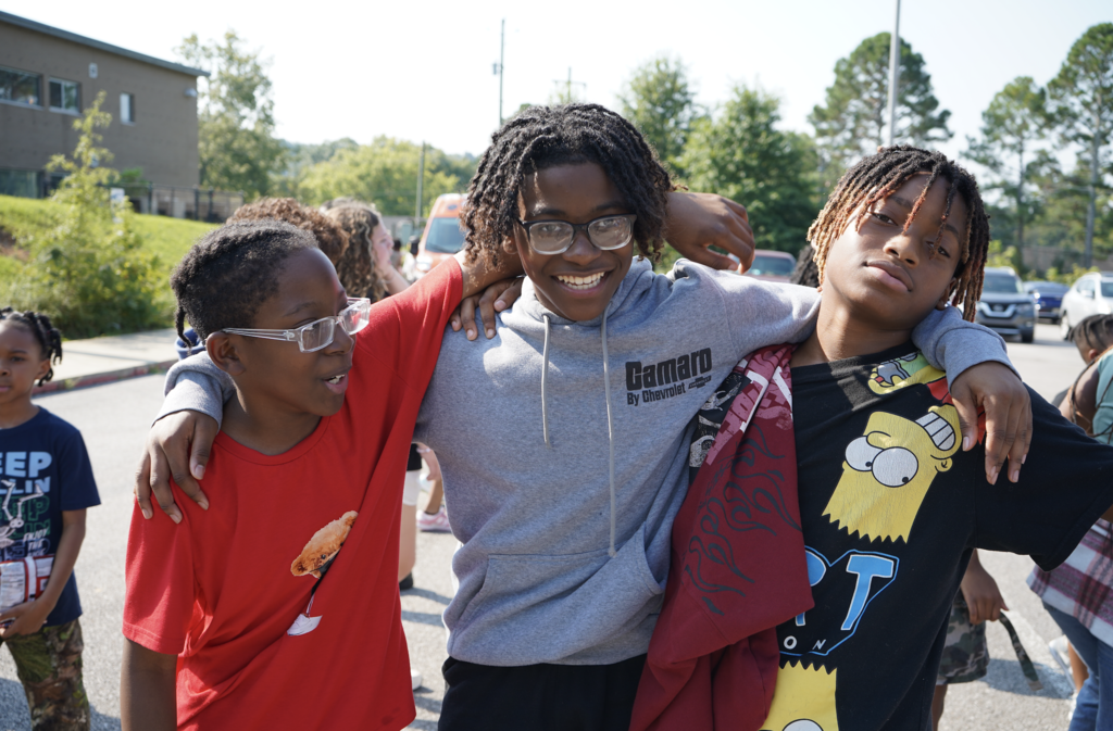 Three students hug each other and smile while standing outside.