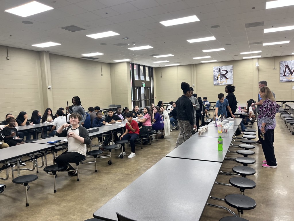 Students and adults sit and stand in a lunch room while eating ice cream.