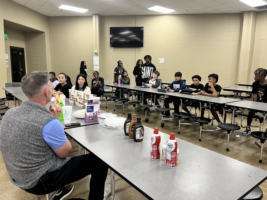 A group of students and a principal sit in a lunch room. Ice cream and toppings are on top of the cafeteria tables.