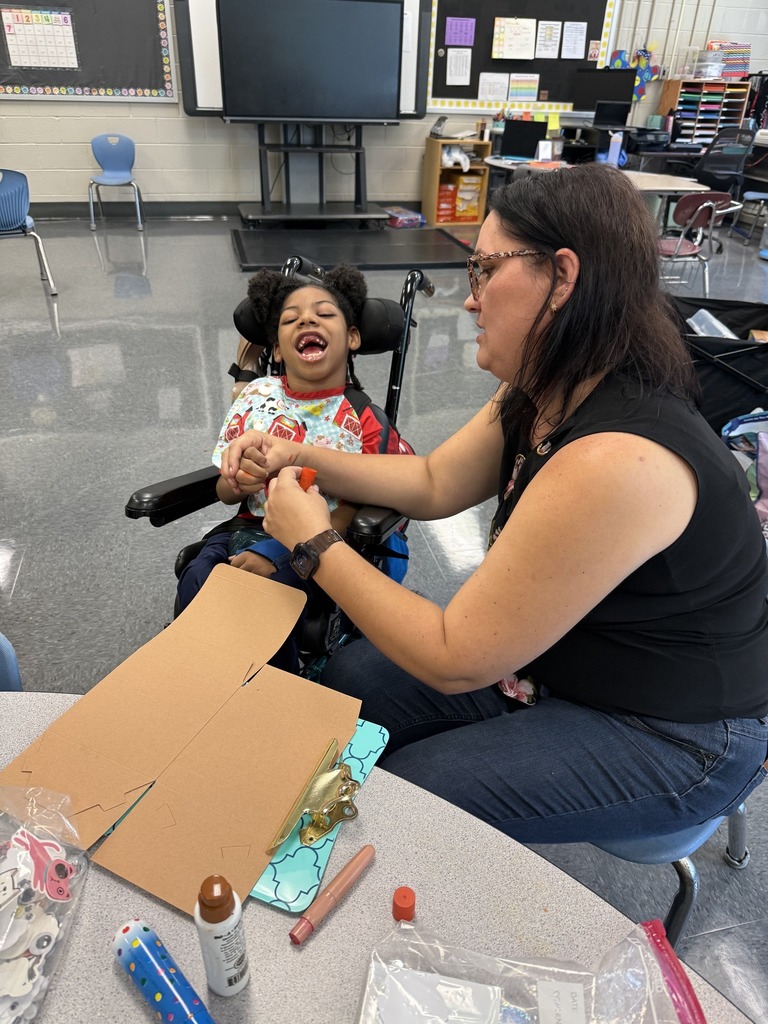 A woman and a young student sit together in a classroom. The woman is sitting in a chair and the student sits next to her in a wheelchair. The woman is helping the student with an arts and crafts project.