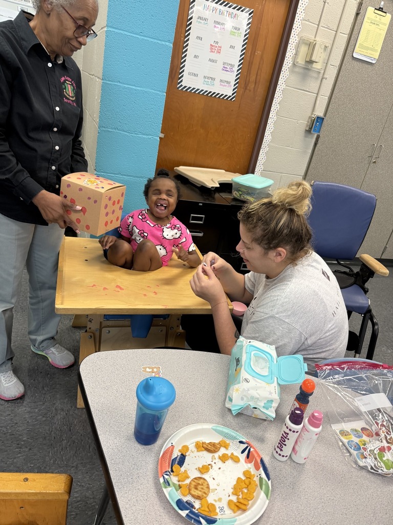 A student sits at a desk while two adults help her with an arts and crafts project.