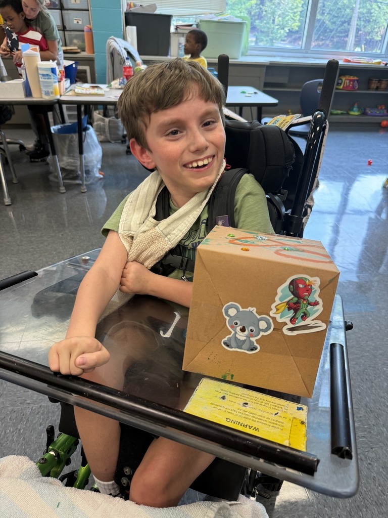 A student sits in a classroom and smiles. The student is in a wheelchair with a desk attached to it. A box with stickers sits on the desk attachment.