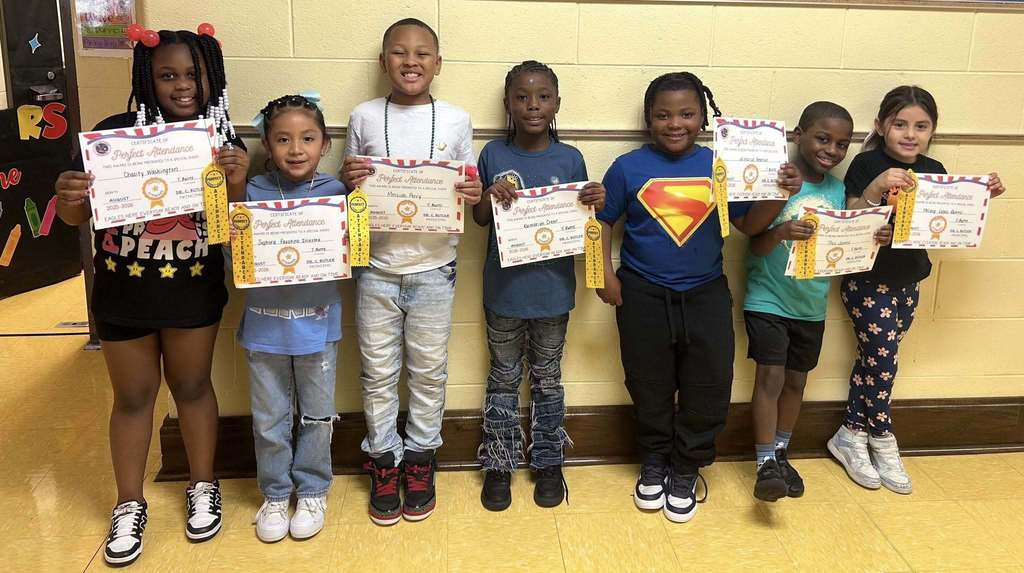 A group of students stand in a school hallway, holding up certificates and ribbons.