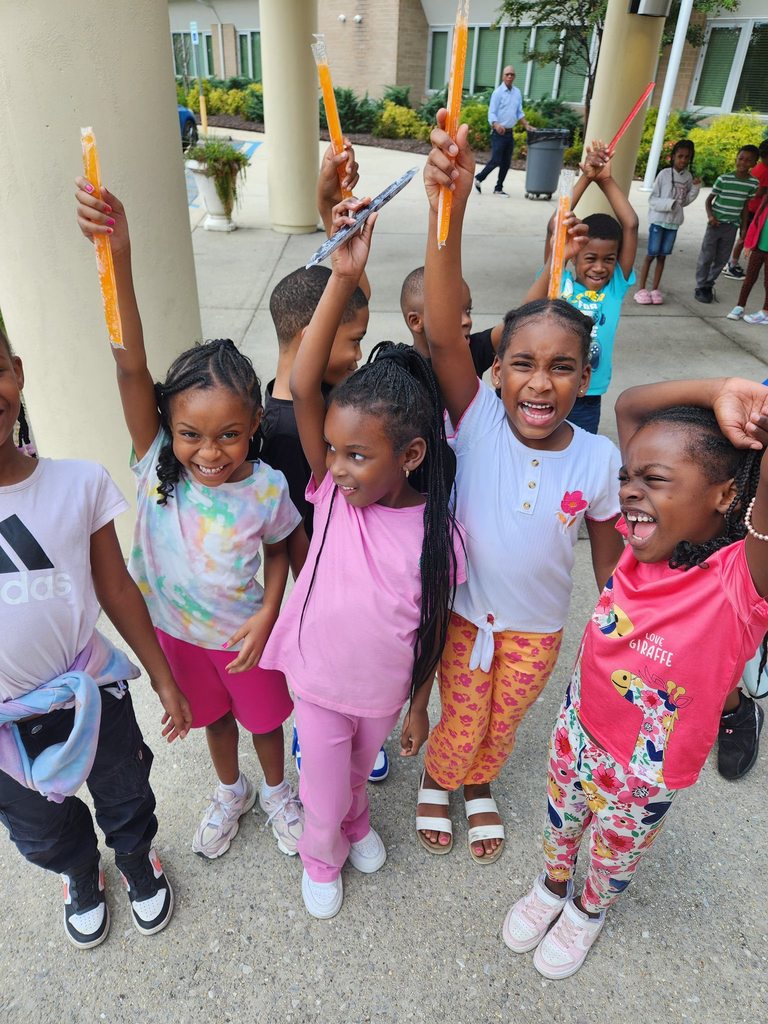 A group of students stand outside a school, holding up popsicles.