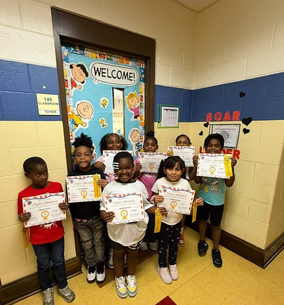 Eight students stand in an elementary school hallway, holding certificates and ribbons.