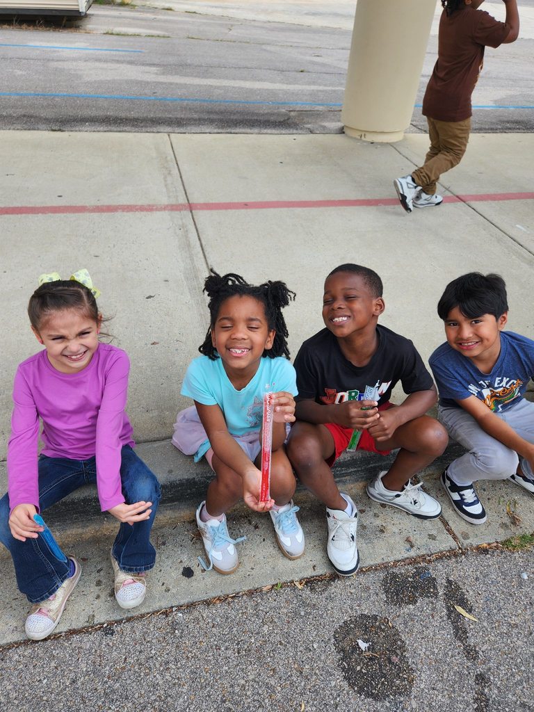 A group of students sit on a sidewalk outside a school while eating popsicles.