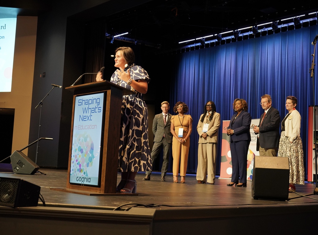 A woman speaks into a microphone at a podium on a stage. A group of adults stand behind her and smile. 