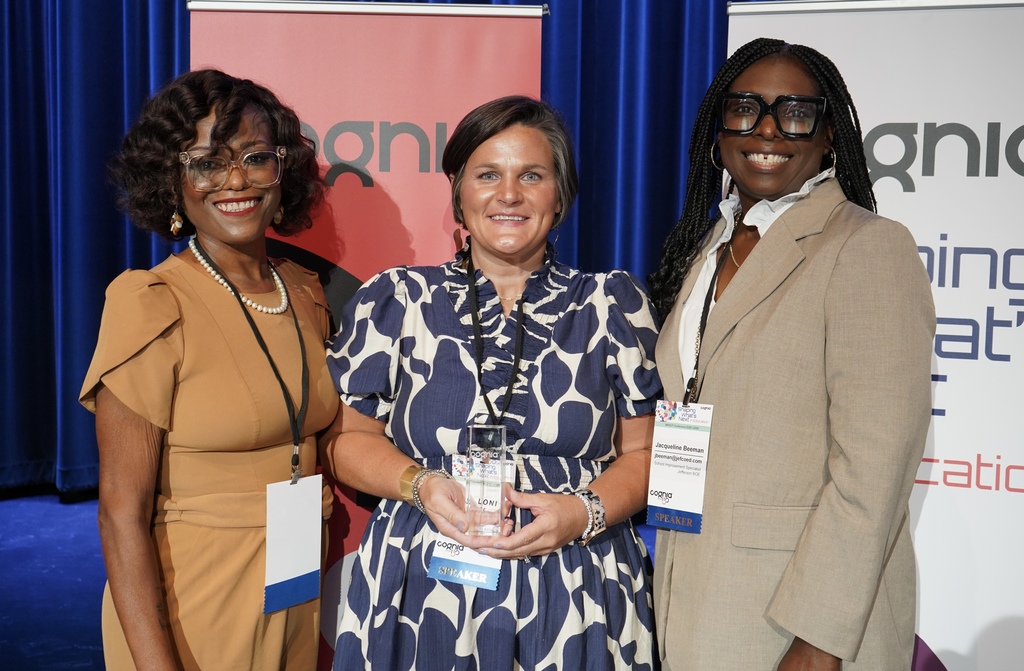 Three women stand on a stage and smile. One of the women holds an award. 