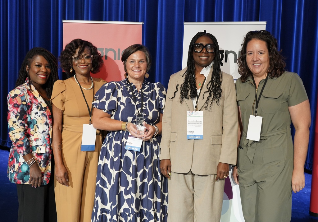 Five women stand together on a stage and smile. One woman holds an award. 