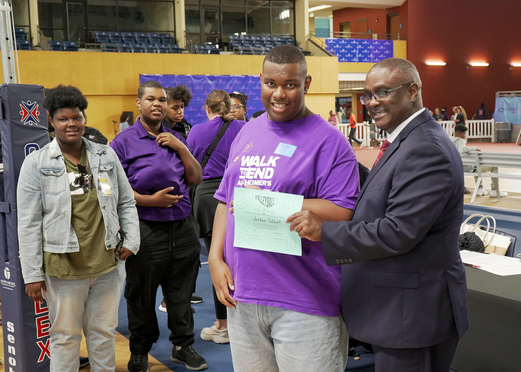 A male student in a bright purple shirt receives a certificate following his participation in and adaptive volleyball event. 