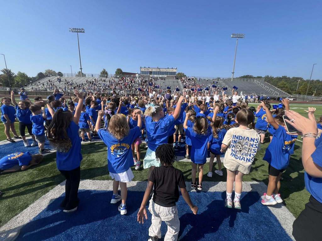 Kindergarten students stand on a football field, watching high school cheerleaders perform.