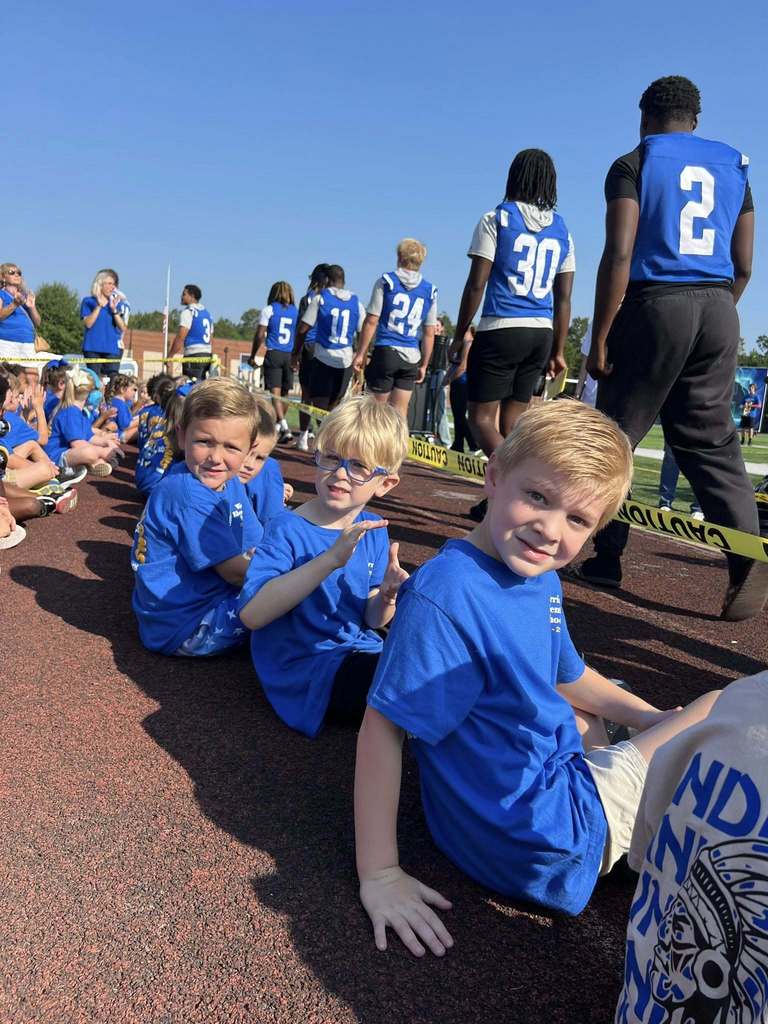 A group of little boys sit on an outdoor track as football players walk past them.