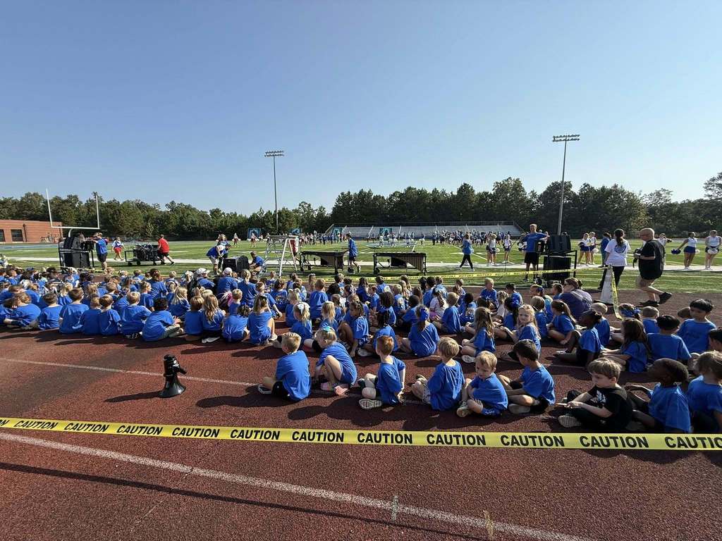 Students sit on an outdoor track, watching high schoolers perform at a pep rally on the football field.