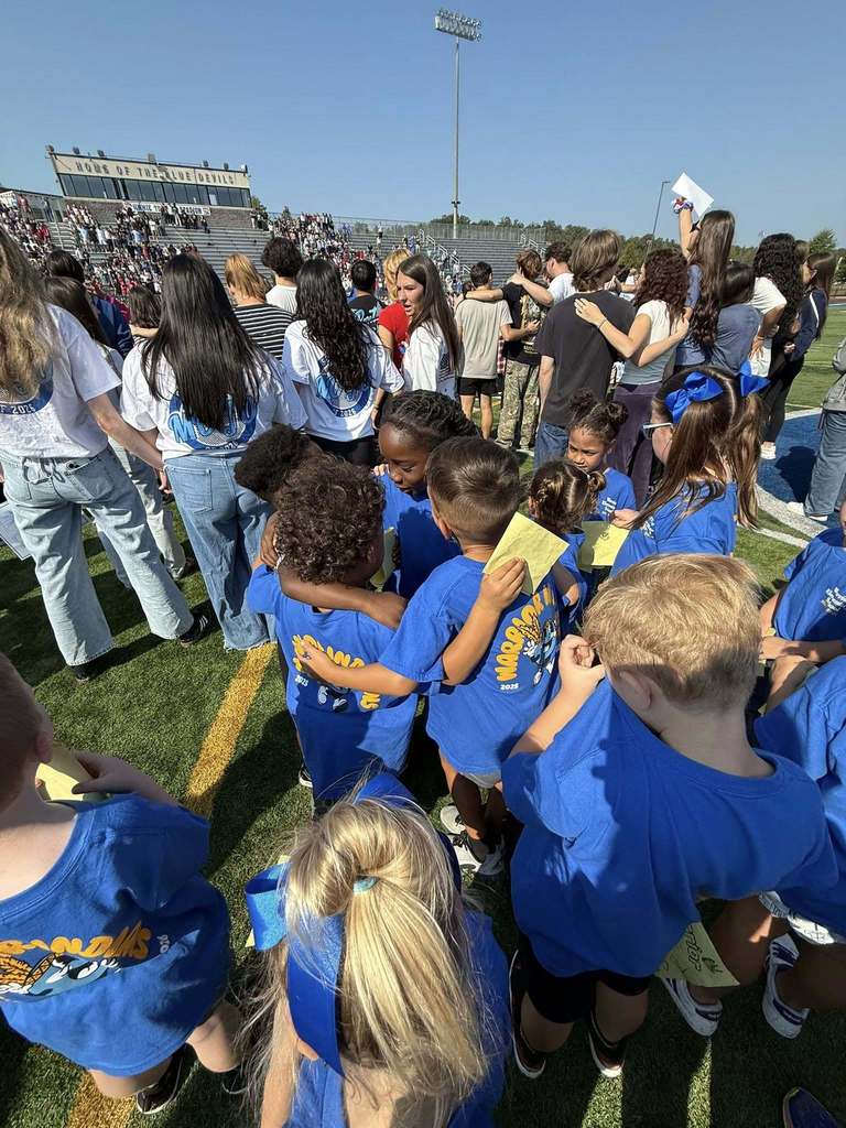 Kindergartners and high school seniors stand together on a football field, holding letters. A group of kindergartners stand in the center of the photo, hugging.