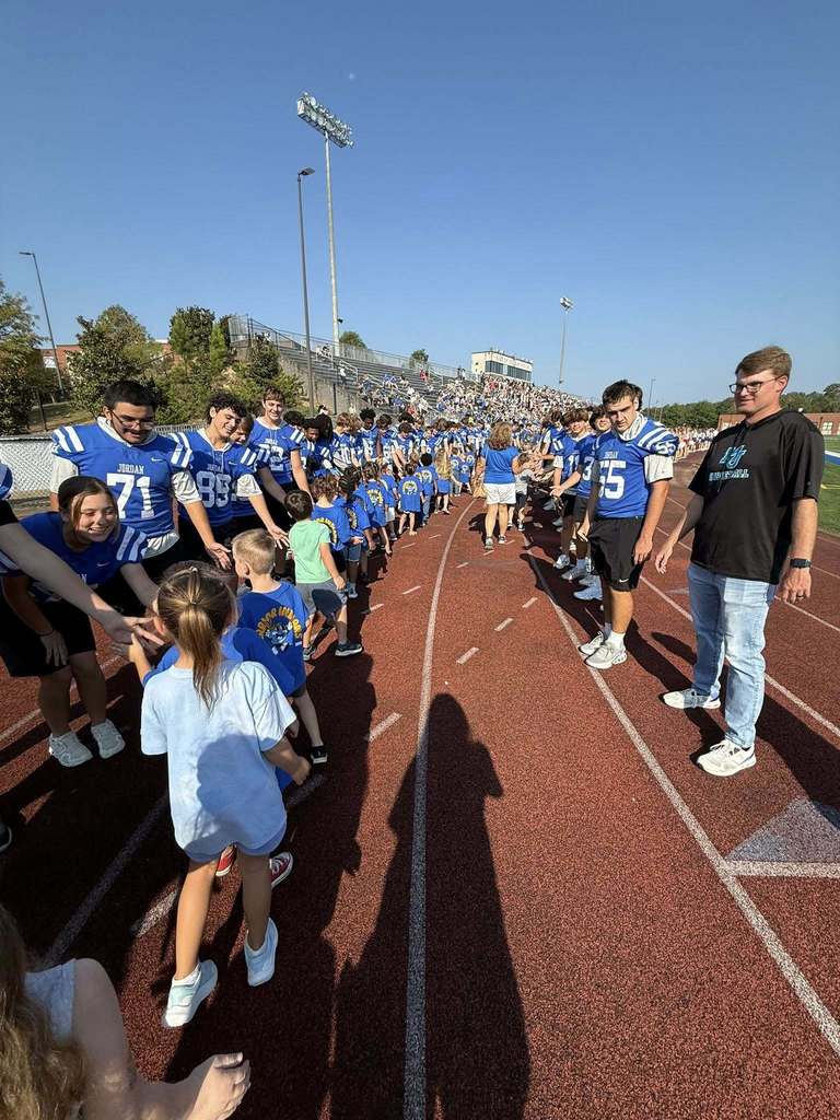 Kindergartners walk along an outdoor track, high-fiving football players.