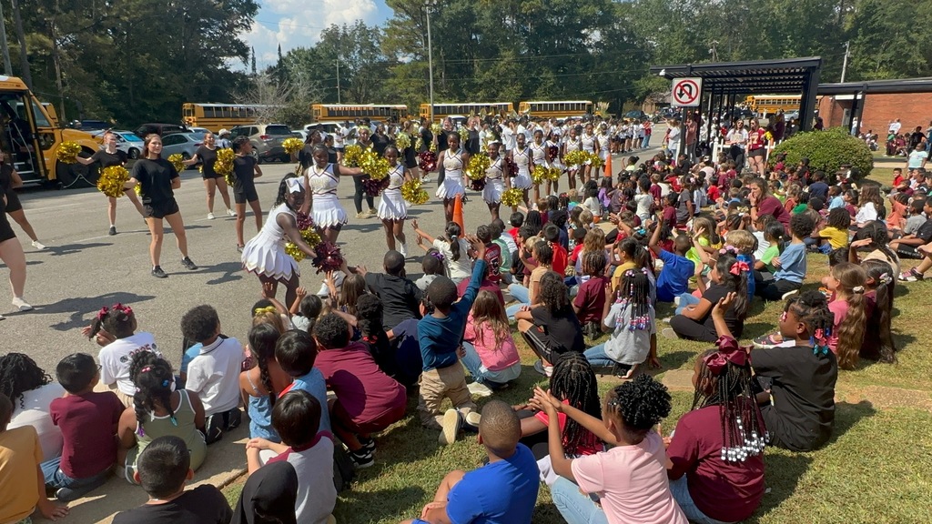 Elementary school students sit in the grass and watch cheerleaders and band members perform in a school parking lot.
