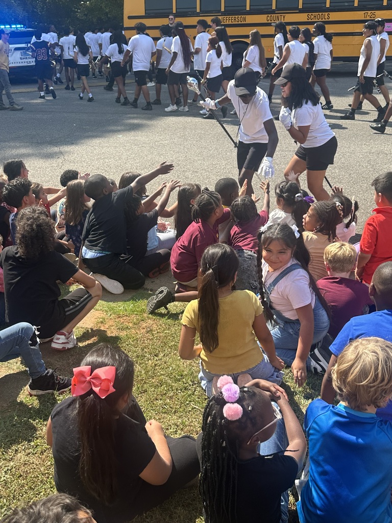 Drum majors for a high school marching band high five elementary school students as they sit in the grass.