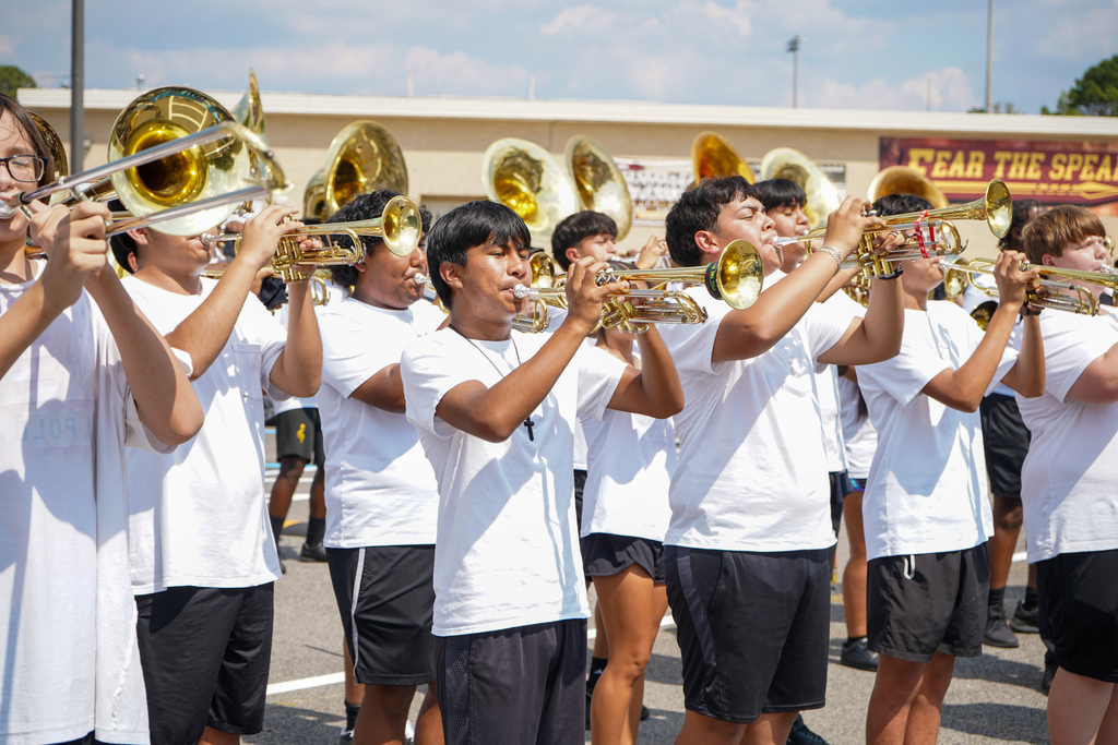 Marching band students wearing white shirts and black shorts perform in a school parking lot