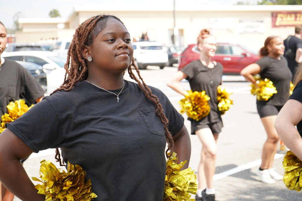 Students wearing matching black shirts and shorts hold pom poms while standing in a parking lot.