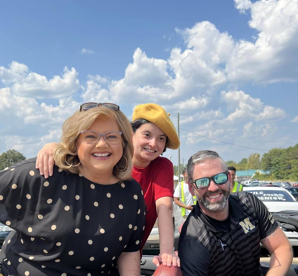 Two women and a man sit in a car outside and smile at the camera.
