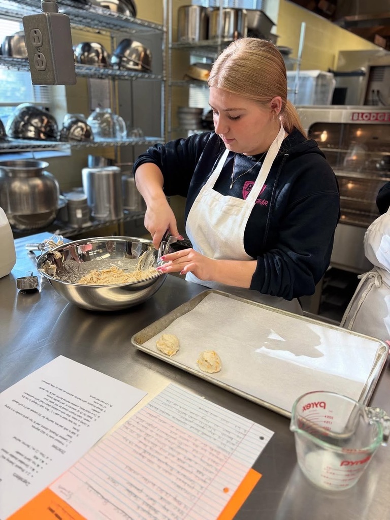 A student scoops biscuit dough onto a pan while standing inside a culinary class kitchen.
