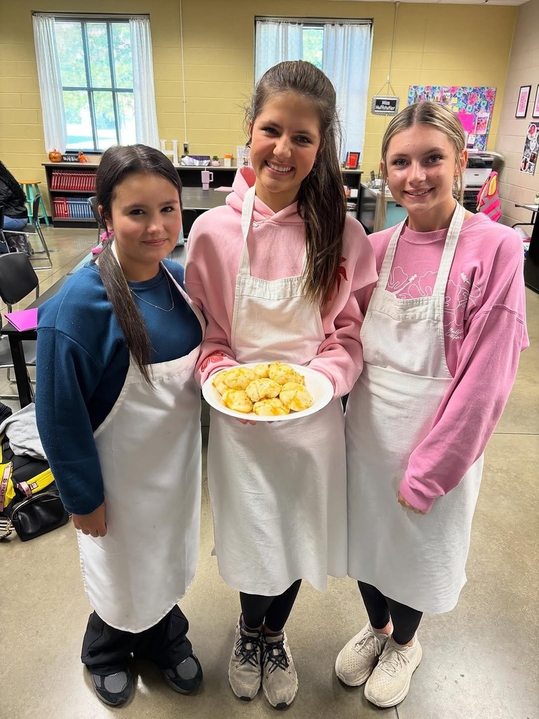 Three students stand together inside a classroom and smile while holding a plate of garlic cheese biscuits