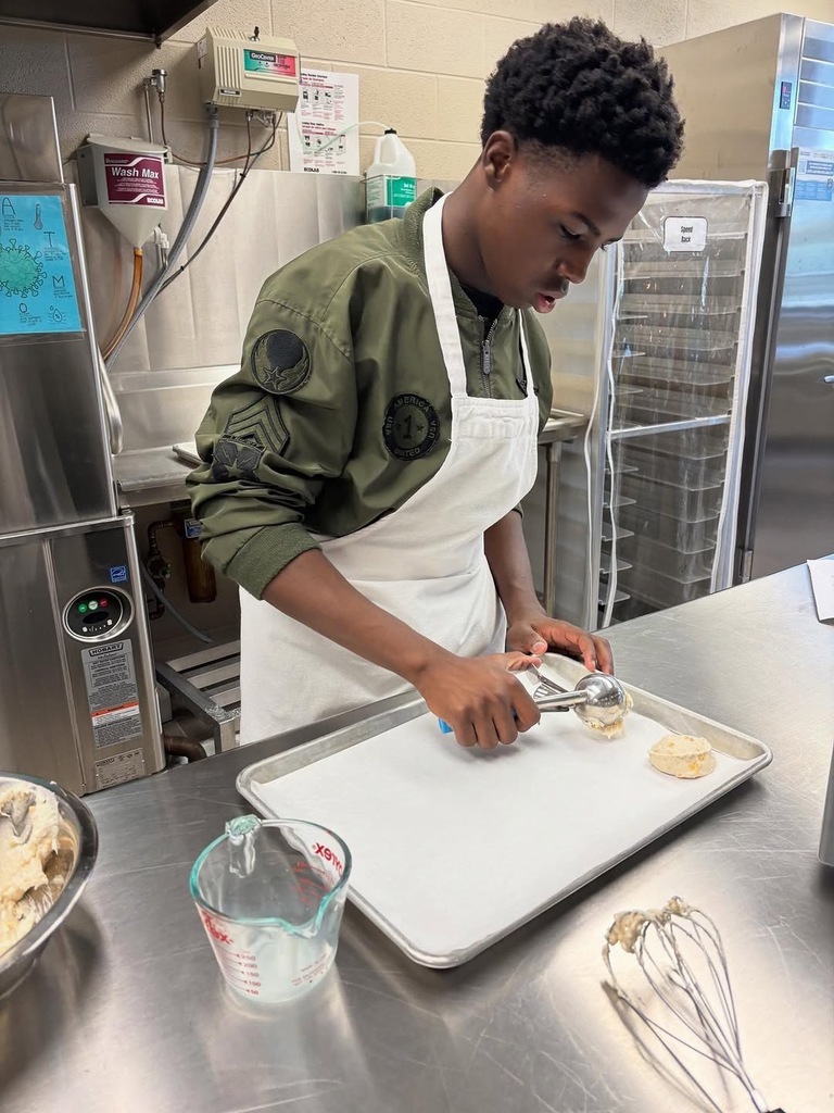 A student scoops biscuit dough onto a pan while standing inside a culinary class kitchen.