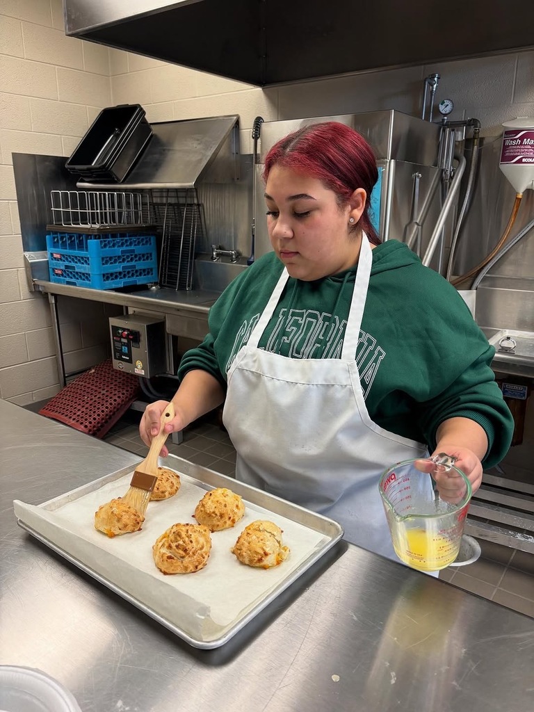 A student brushes butter onto garlic cheese biscuits while standing inside a school's culinary class kitchen.
