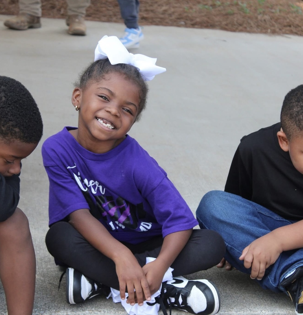 A little girl with a bow in her hair sits on a sidewalk and smiles. 