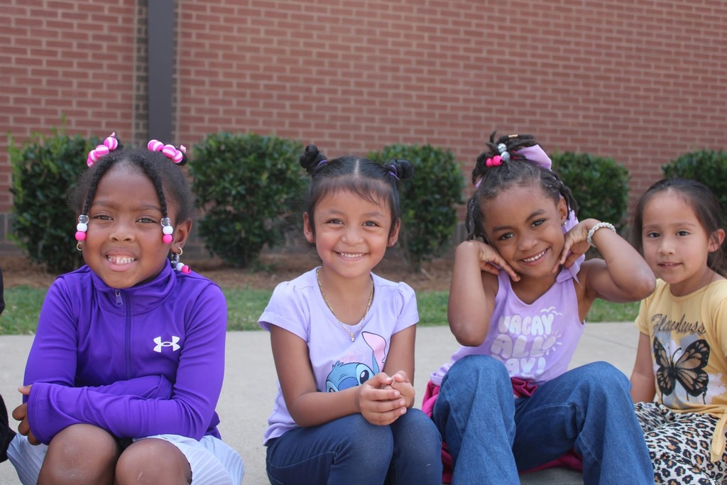 Four elementary school students sit on a sidewalk and smile.