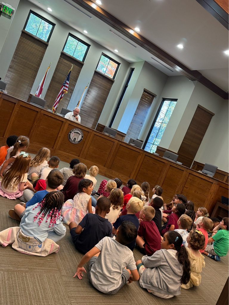 A group of kindergarten students sit on the floor of the Gardendale City Hall chamber. The mayor sits in front of them, speaking into a microphone.