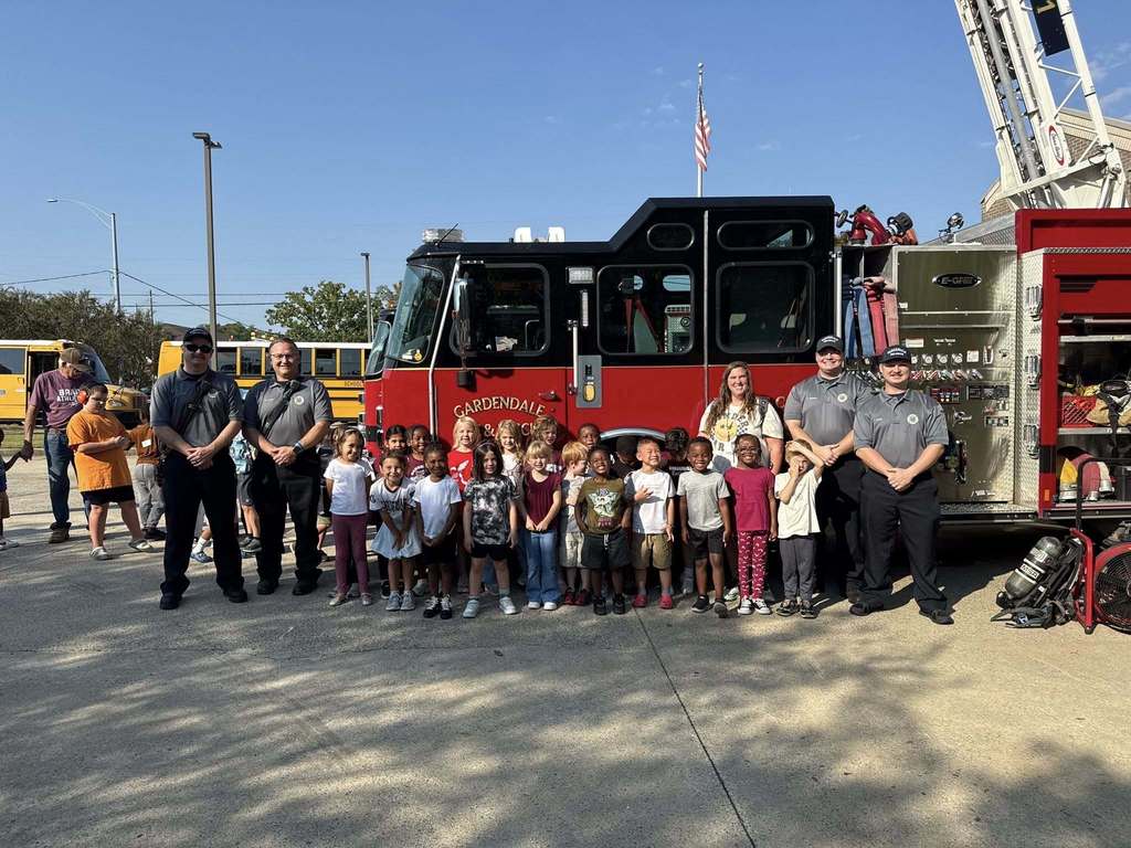 A group of kindergarten students, firefighters and two other adults stand in front of a fire truck.