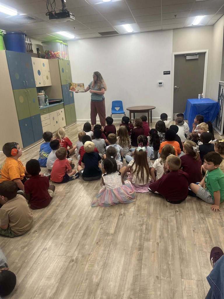 A woman stands and reads a book to a group of children who sit on the floor.